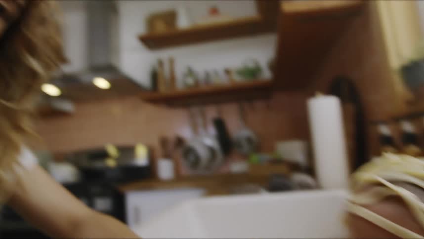 A woman laughs at her infant sitting in a sink playing with pasta