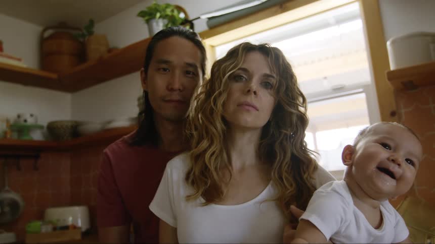 Father with long hair and mother with brown and wavy hair holds baby in the kitchen looking at camera