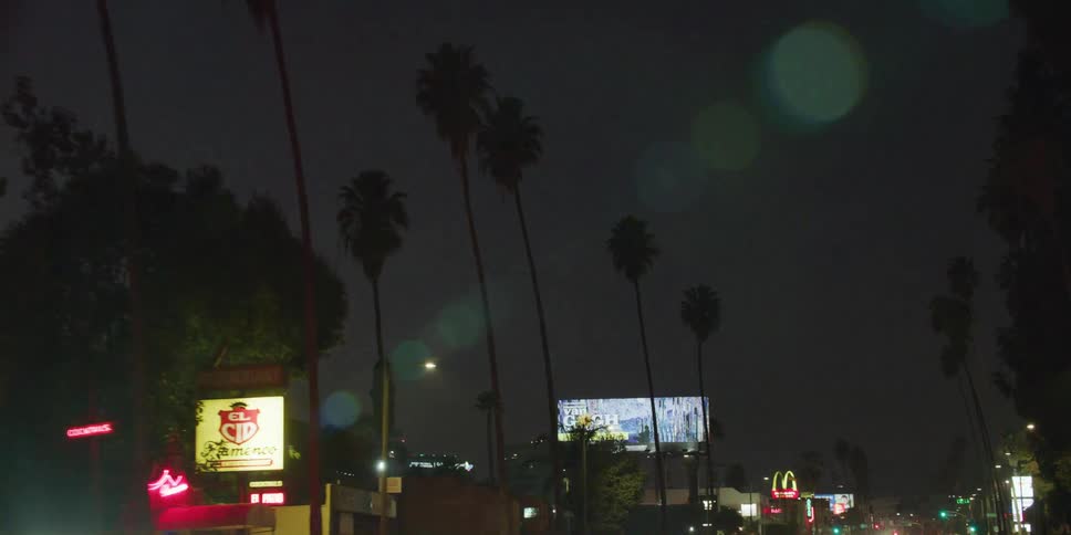 Long stream of cars with their headlights on drive along street past businesses at night