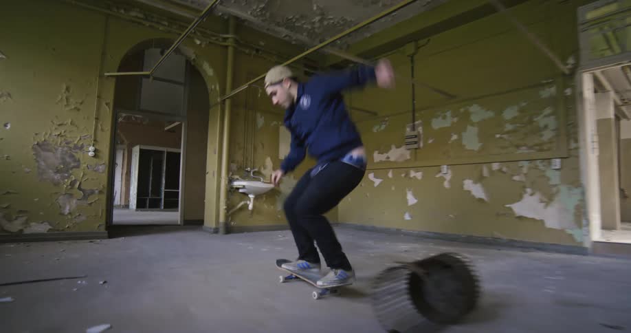 Man rides a skateboard in an empty building and leaps over a metal basket with it