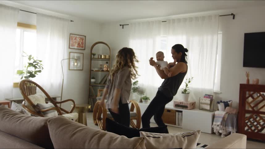 Father with long hair undershirt and black pants holds baby son dancing with mother with brown wavy hair in living room at home
