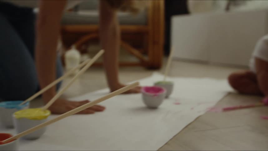 Mom sets paint bowls and brushes on paper rolled out on the floor and tries to show baby how to finger paint