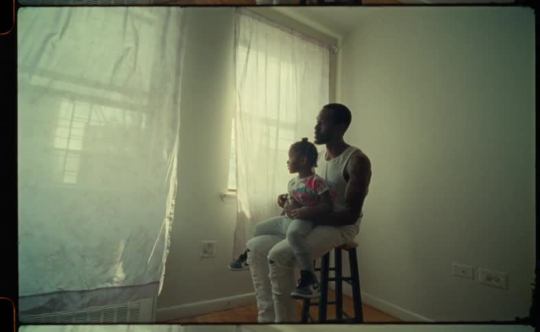 Man sits on a stool and holds his daughter on his lap in an otherwise empty room as curtains in the breeze