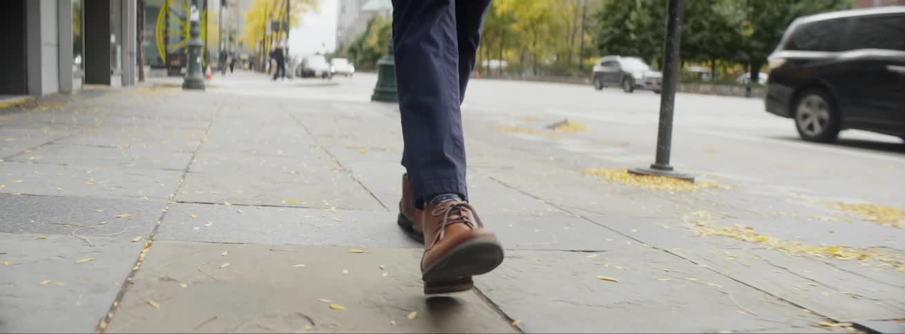 Businessman with navy blue pants and brown leather shoes walks down the street on the pavement next to highway with cars driving