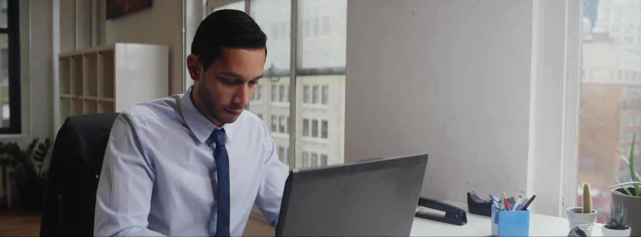A man is sitting at a desk in an office typing on a laptop computer