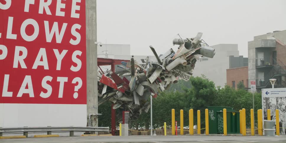 Sculpture of transport pieces stands near bulletin board and apartment buildings