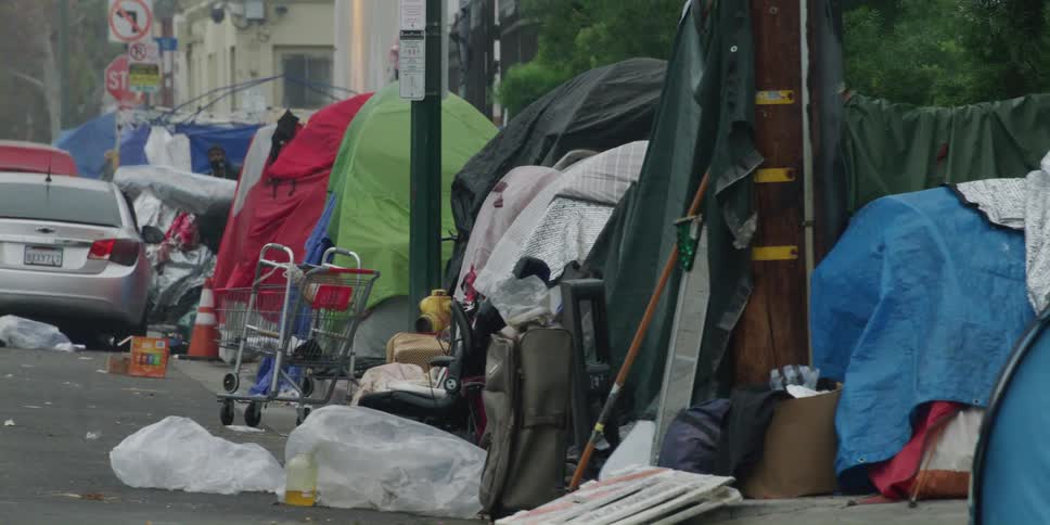 Mangled tents and debris litter the ground and sidewalk in a homeless camp