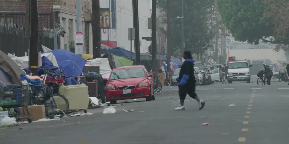 A man crosses the street toward a homeless camp of tents and tarps on a city sidewalk