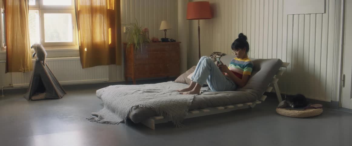 Girl with stripped shirt and ripped jeans sits on bed using smartphone in bedroom