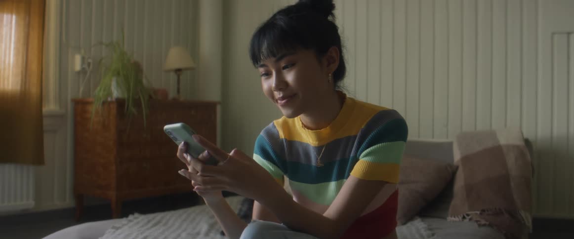 Girl with glasses earrings and stripped tshirt sitting on bed uses smarphone in bedroom