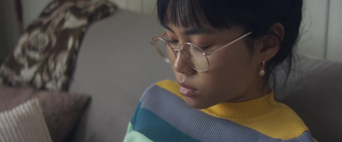 Girl with glasses earrings and stripped shirt sitting on bed uses smartphone in bedroom