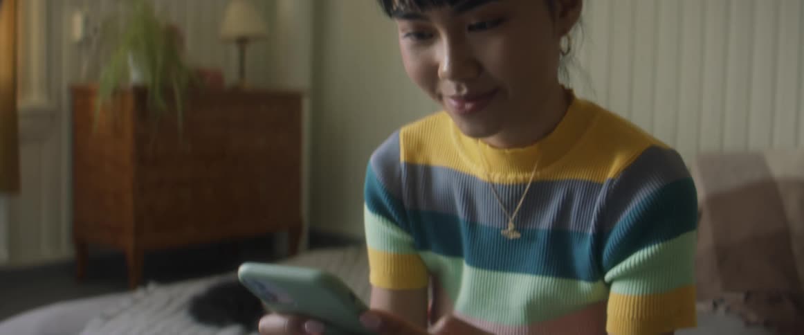 Girl with stripped shirt and ripped jeans sits on bed using smartphone in bedroom