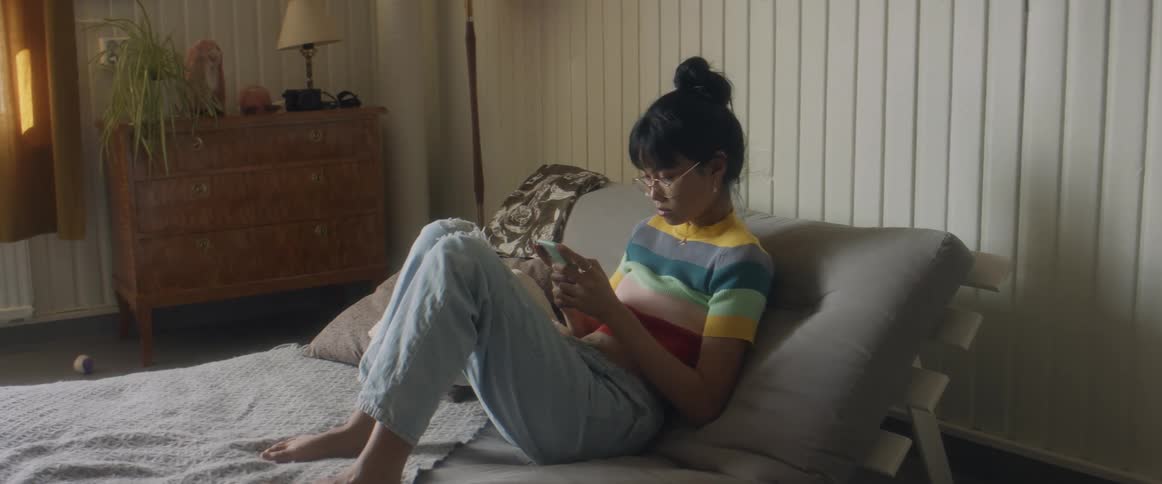 Girl with glasses stripped shirt and ripped jeans sits on bed using smartphone in bedroom