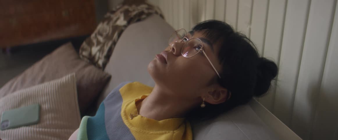 Girl with glasses and stripped tshirt sitting on bed puts smartphone down looking up at ceiling in bedroom