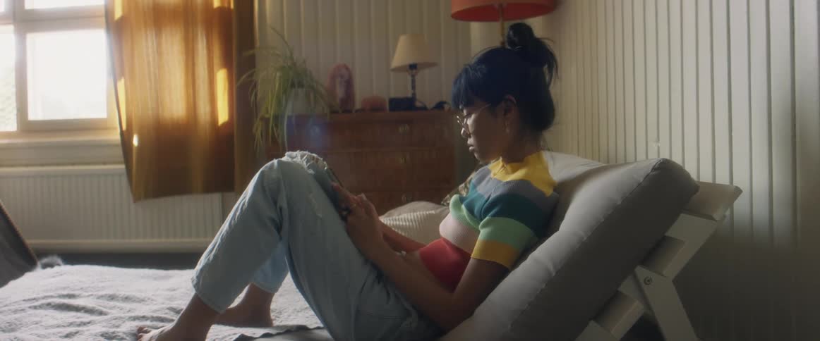 Girl with glasses stripped tshirt jeans sitting on bed uses smartphone in bedroom