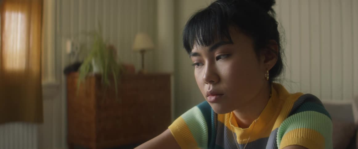 Asian woman sits thinking on a bed with a table and lamp and plant nearby