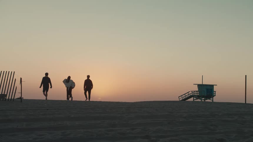 A young woman and two men walk towards the sunset on a beach