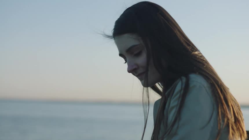 Woman with straight hair and overcoat looks at sea on the coast at sunset