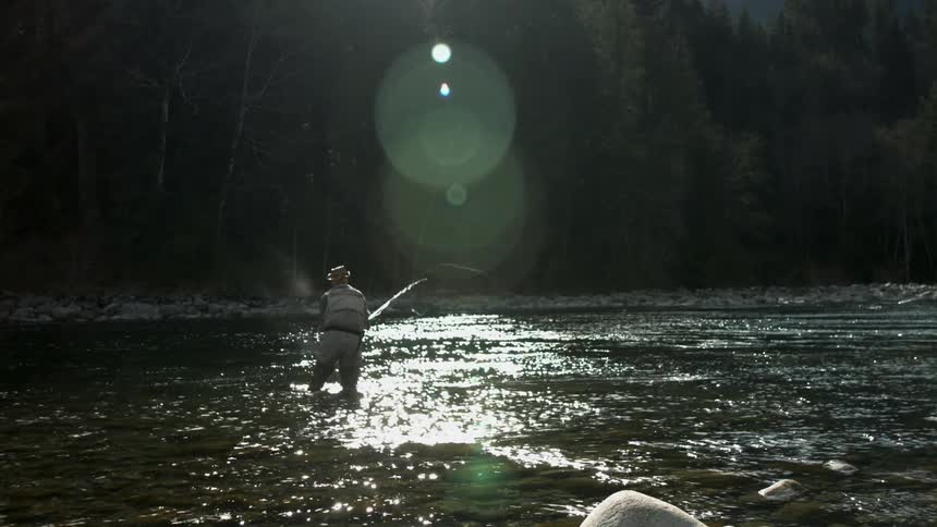 A man casts a fishing line back and forth while standing in a river