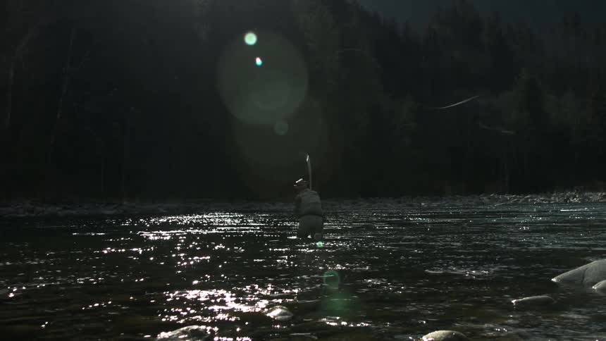 A fisherman standing in a river casting his line as the water flows and shimmers around him