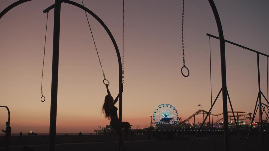 The silhouette of a woman twirling and swinging on the rings at the santa monica pier with ferris wheel shimmering behind