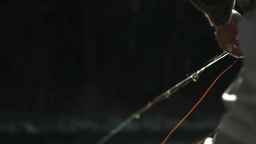 Fishing man adjusts fishing line while standing on bank across water from forest