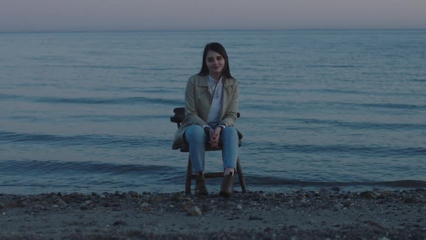 A teenage girl is sitting in a chair on a beach as the waves roll in under her