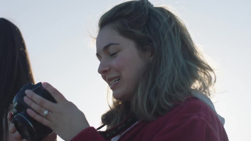 Young white women wearing coats and standing outdoors while one of them takes pictures with a photographic camera and the other one holds a mirror