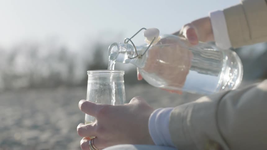 Woman sits on beach pouring clear liquid from bottle into a glass garnished with a lemon slice