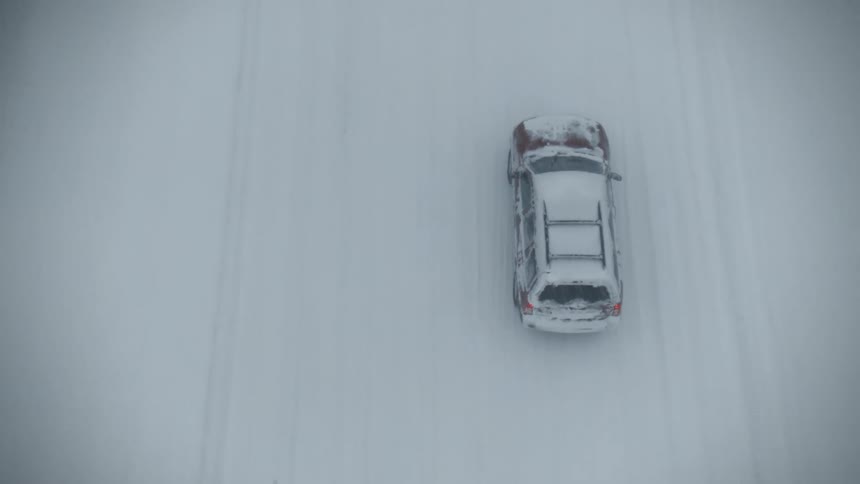 Car with hazard lights on drives on snowy road while snowing