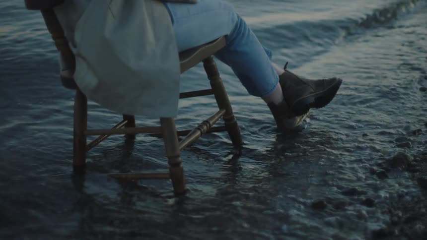 A woman sits on a wooden chair as the tide roles to the shore