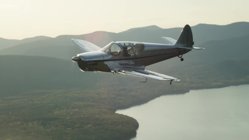 The sun is shining brightly upon a small airplane flying over a forest and lake with mountains in the distance