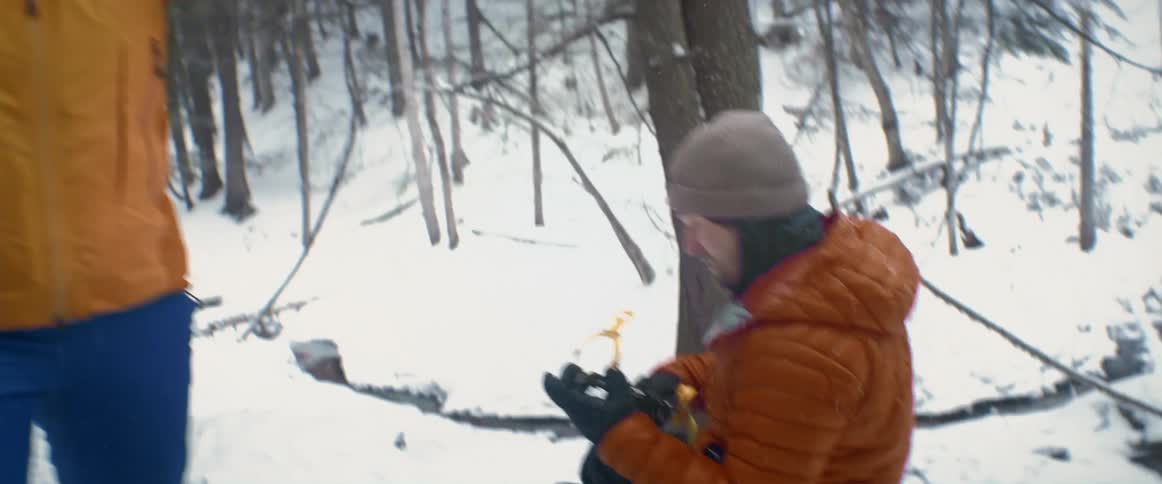 A man sits in the snow and looks at the ice picks for his shoes while a woman standing next to him puts her helmet on