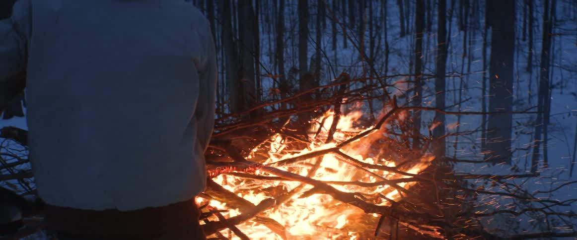 A man is putting more wood on a large bonfire in the woods on a winter evening