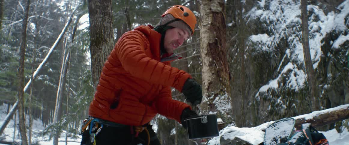 A man is cooking out in a snowy area as a woman can be seen sitting nearby