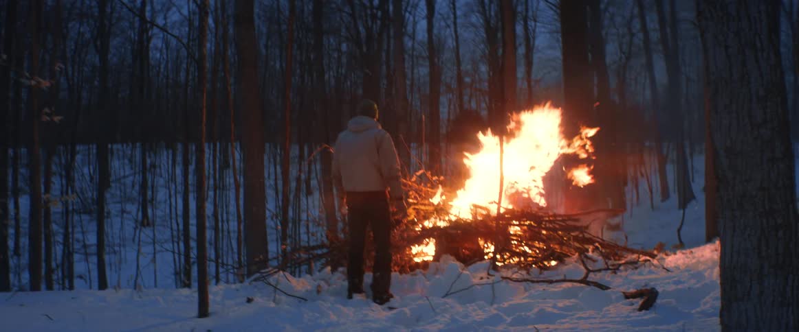 A man is building a large bonfire in the woods on a winter evening