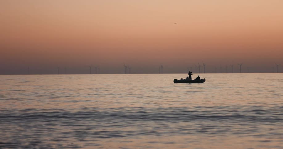 A person stands on a boat and looks towards wind turbines in the distance while another person sits on the boat