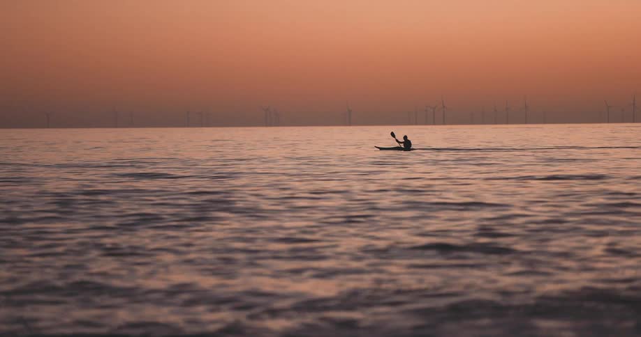 Silhouetted person paddles across waves under a red sky as windfarm turbines turn slowly on the horizon