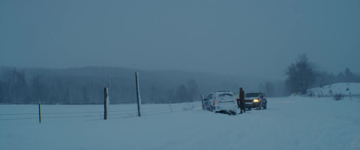 Man walks around a vehicle stuck in the snow near a fence on a rural road at winter