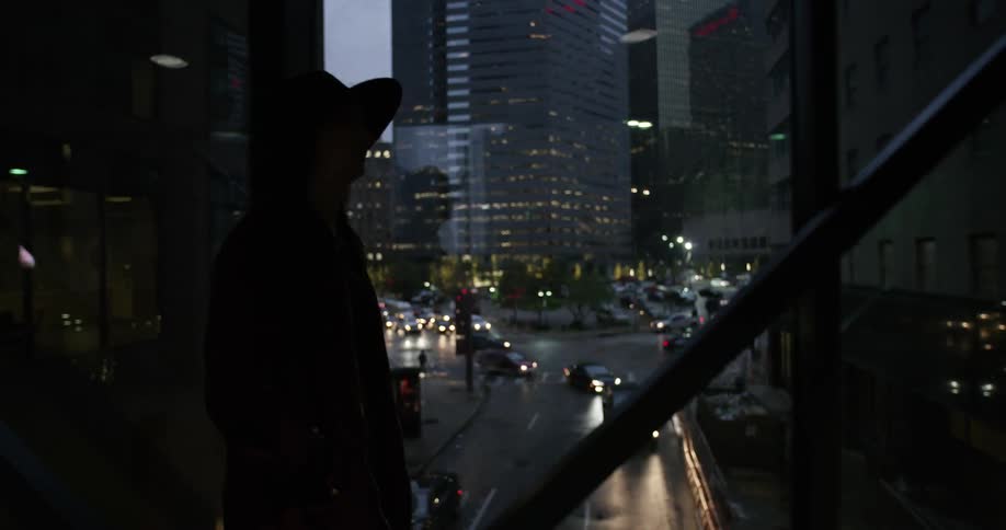 A woman in a black hat walks down a darkened glass hallway