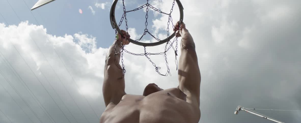 Shirtless young black man doing pull ups in the basketball hoop outdoors