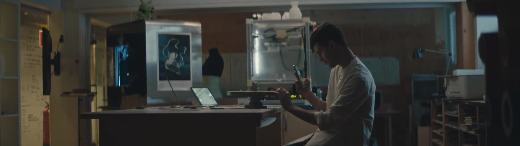 A man is sitting at a desk with a laptop computer and taking a picture with his phone of piece of iron pyrite in front of him