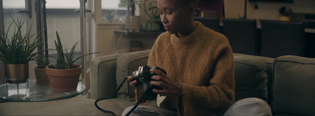 Young black woman wearing a sweater sitting at a couch handling her photographic camera