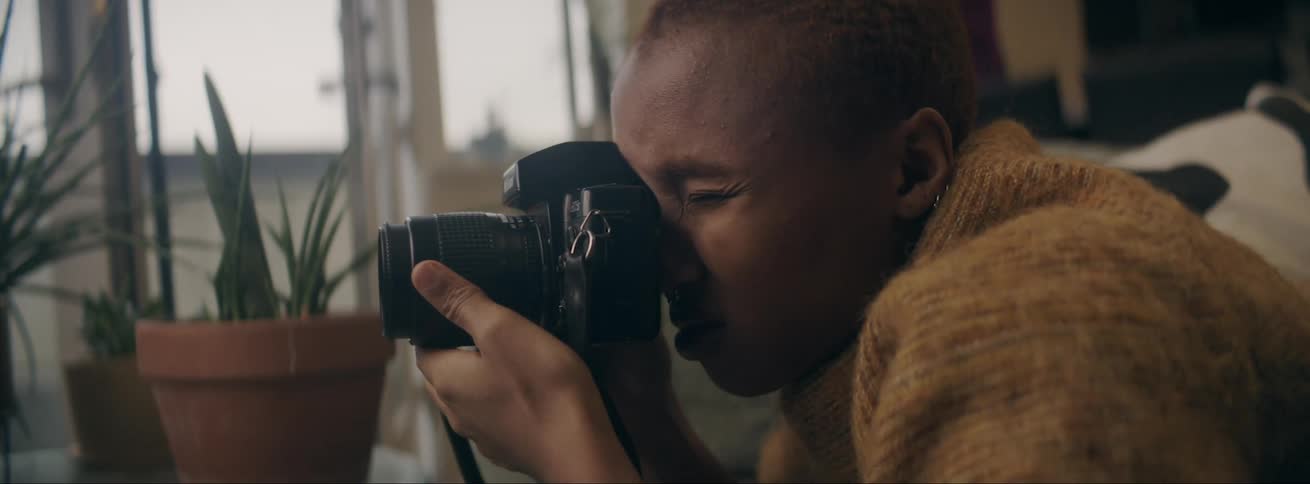 Black young woman wearing a yellow sweater at her living room using her photographic camera to take pictures
