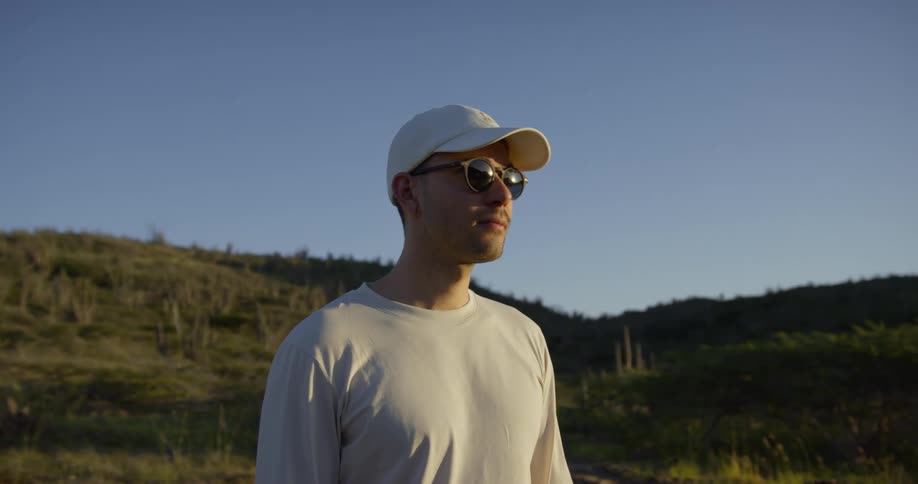 White man wearing a white cap and sunglasses standing in the middle of a hill covered in grass and plants