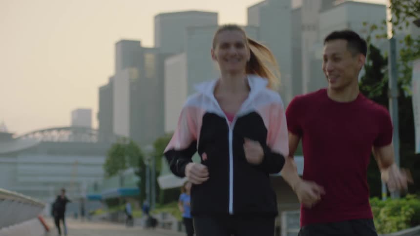 Asian man and white woman jogging together at a public space in a big city with buildings and skyscrapers at the background