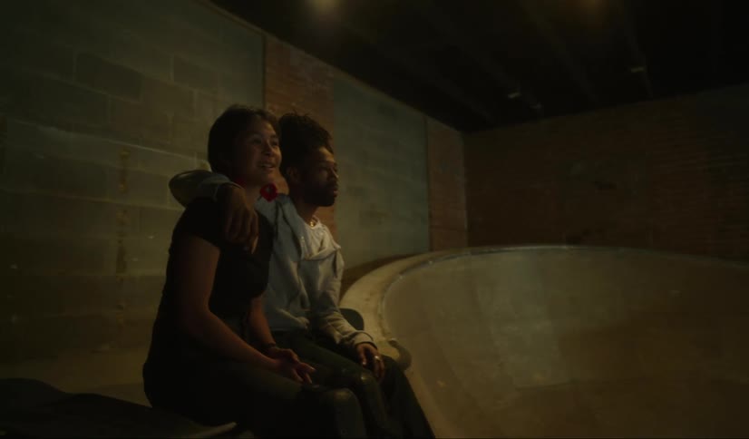 Multi ethnic couple sit and embrace watching others skateboard at an indoor covered skate park