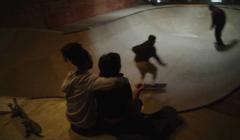 Two men skating at an indoor skatepark while a man and a woman are sitting nearby watching them