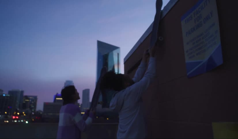 Two teenagers set their skateboards on a rooftop as they climb on top of a tower on a building rooftop