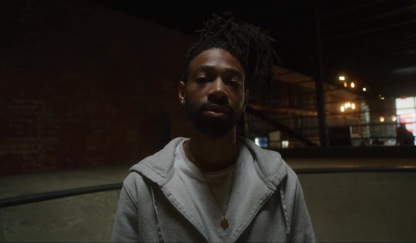 Black man wearing a grey hoodie stading alone in the middle of an indoor skatepark